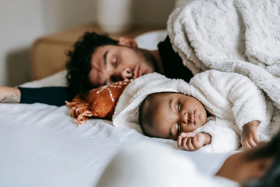 Père endormi à côté de son nouveau-né sur un lit, couvertures claires